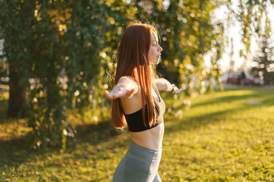 Side view of relaxed redhead yogini young woman with closed eyes standing with hands divorced to side and deeply breathing, outside in city park background of bright sunray in evening at sunset. - Powered by Adobe