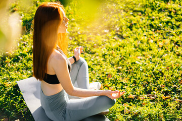 Side view of young red-haired woman with closed eyes meditating in lotus pose holding hands on knee, sitting in yoga mat on green grass at city park in sunny summer morning.