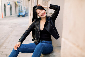 Obraz na płótnie Canvas Beautiful sexy young Asian woman with ponytail hair, wearing trendy casual clothes, leather jacket and denim jeans, sitting outdoors on stairs in the city, smiling to camera
