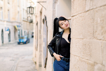 Front view of young charming brunette Asian woman in trendy clothes, posing at the street city against the background of the wall of old building