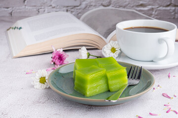 Layer sweet cake (or khanom Chan) on a plate and a cup of white coffee with a book and flowers on gray stone background. Space for text. Selective focus. Relax time. Concept of relax time