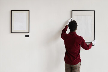 Back view at African-American man hanging blank frames on wall while planning art gallery or...