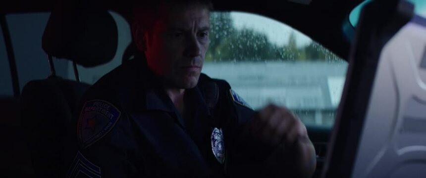 HANDHELD Portrait of police officer talking on CB radio while checking information on a laptop inside a car. Shot on RED Dragon with 2x Anamorphic lens