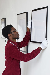 Fototapeta premium Vertical side view at African-American man hanging frames on wall while planning art gallery or exhibition