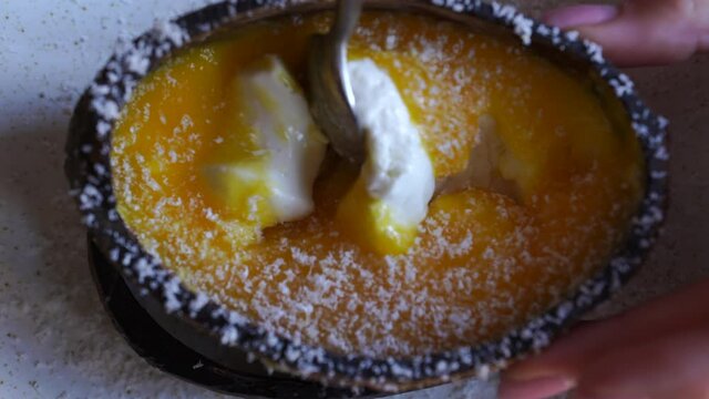 With a metal teaspoon, the girl is stirring yellow mango with pannacotta, sprinkled with coconut flakes, coconut shell. Delicious evening dessert in a restaurant, cafe. Social media for business