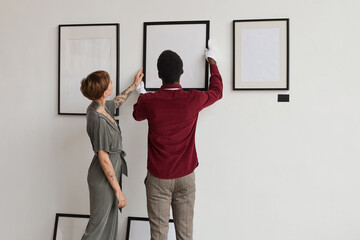 Back view portrait of two art gallery workers hanging painting frames on white wall while planning...