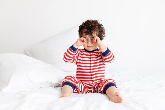 Sleepy toddler wearing pajamas sitting on white bed rubbing eyes
