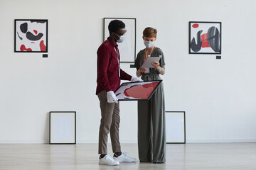 Full length portrait of creative tattooed woman looking at paintings with African-American man while planning art gallery exhibition, copy space