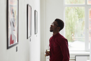 Side view portrait of young African-American man looking at paintings and holding champagne glass...