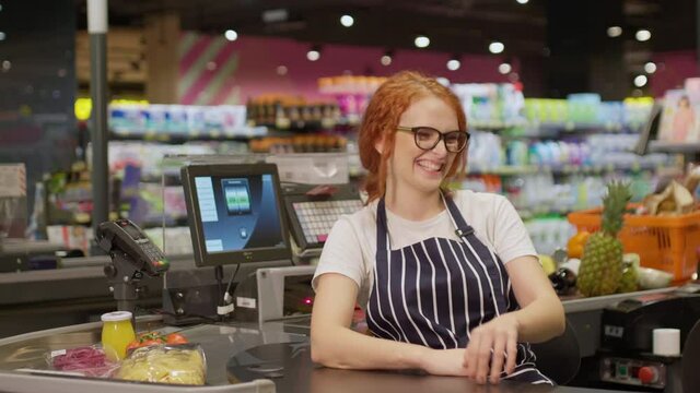 Pretty cheerful redhead cashier in eyeglasses and striped apron happily posing on camera working behind cash box in modern supermarket. Grocery store concept