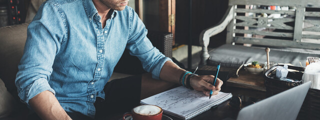 Young entrepreneur working on business project in city cafe, writing down ideas and using laptop....