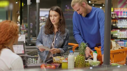 Young attractive couple with checklist laying out goods at the checkout in modern grocery store. Young family on shopping in supermarket