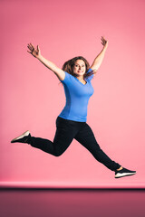 Plus size model in casual clothes jumping in studio on pink background