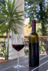 One glass of red wine and an open dark green glass bottle on a wooden table against a blurred landscape background