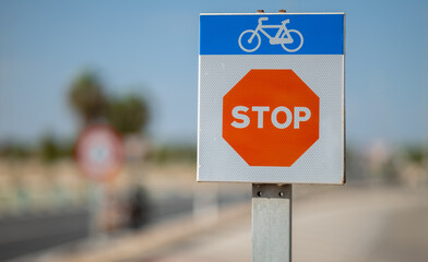 A small stop sign on a bike lane next to a highway. Cyclists have to stop because this is where cars can cross the cycle path. There is a risk of accident. A blue pictogram with a bicycle.