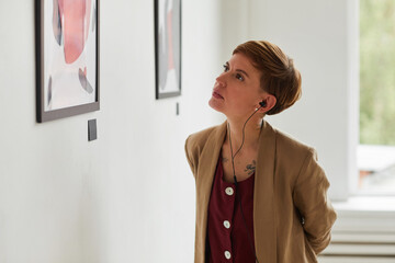 Waist up portrait of tattooed young woman looking at paintings and listening to audio guide at...