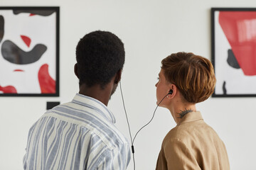 Back view portrait of two young people looking at paintings and sharing audio guide while exploring modern art gallery exhibition, copy space