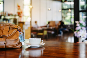 Mini alcohol gel bottle to kill Corona Virus(Covid-19) hang on a brown leather shoulder bag with hot coffee on table in cafe.New normal lifestyle.Health care concept.Selective focus on alcohol gel