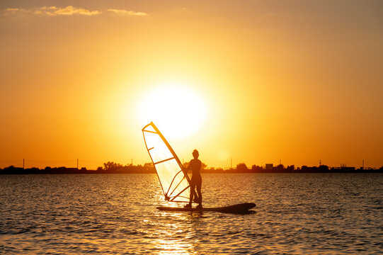 A beginner windsurfer woman stands on a board with a sail on a sunset background. Windsurfing school