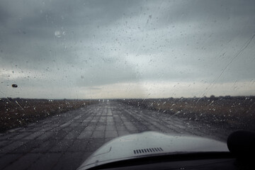 View from the cockpit on the runway in rainy weather. You can see the nose of the plane and raindrops running down the glass. The sky is grey and overcast. High quality photo