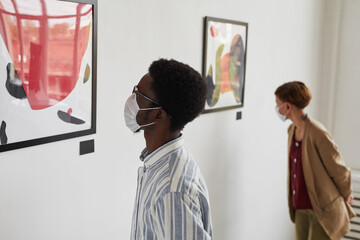 Portrait of two young people looking at paintings while wearing masks at modern art gallery exhibition, copy space