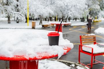 A black paper cup of coffee with a red lid stands in a snowdrift on a table in a street cafe in winter