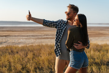 Joyful couple hugging and taking selfie on cellphone on nature