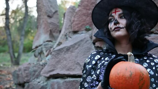 A girl with scary makeup and an evil face holds a pumpkin Theme of costumes and decorations for the Halloween holiday. Traditional makeup for holiday of Santa Muerte