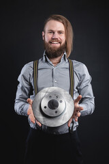 A bearded stylish man in the wheelhouse with suspenders smiles, looking at the camera, and holds a car brake disc in his hand. Ideal auto parts seller © yanik88