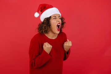 Brunette woman wearing Christmas hat excited and glad to achieve victory, clenches fists, screams in excitement with closed eyes,  successful person.