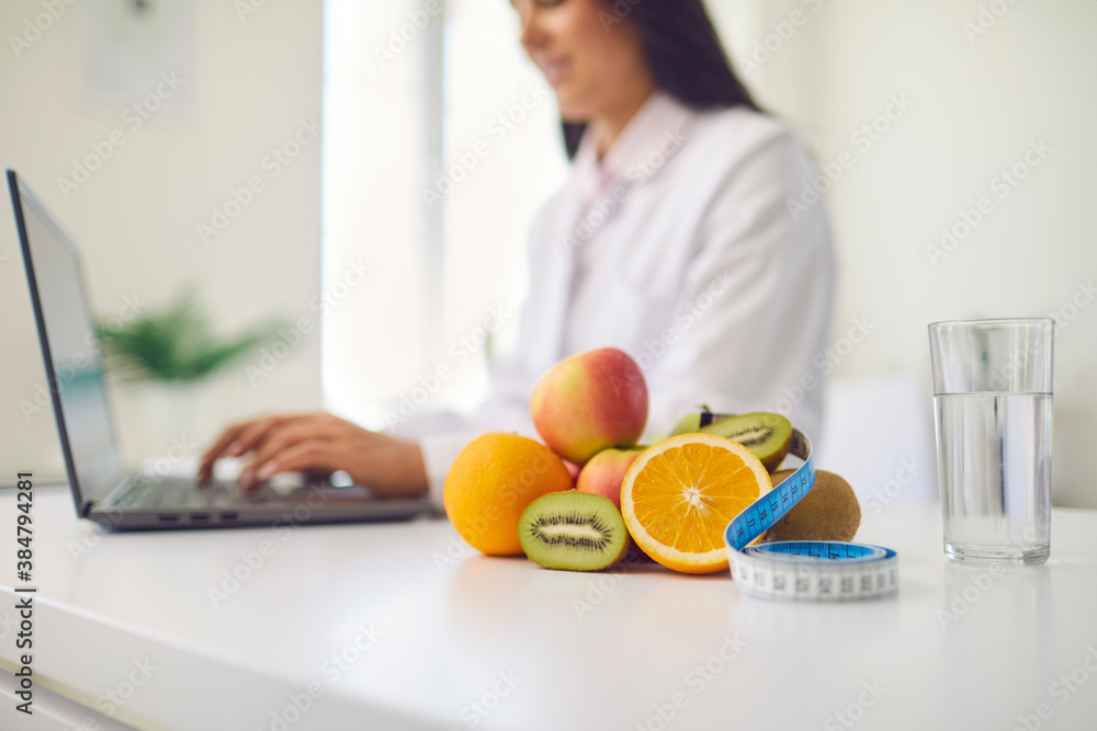 Wall mural fruit, measuring tape and glass of water placed on desk against blurred dietitian working on laptop