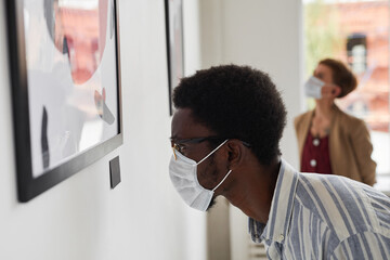 Side view portrait of young African-American man looking at paintings and wearing mask at modern art gallery exhibition, copy space