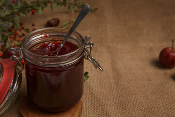  jar of strawberry jam is on a table covered with a rough rustic tablecloth, and next to it lies an apple and a sprig with red berries . selective focus 