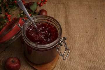  jar of strawberry jam is on a table covered with a rough rustic tablecloth, and next to it lies an apple and a sprig with red berries . selective focus 