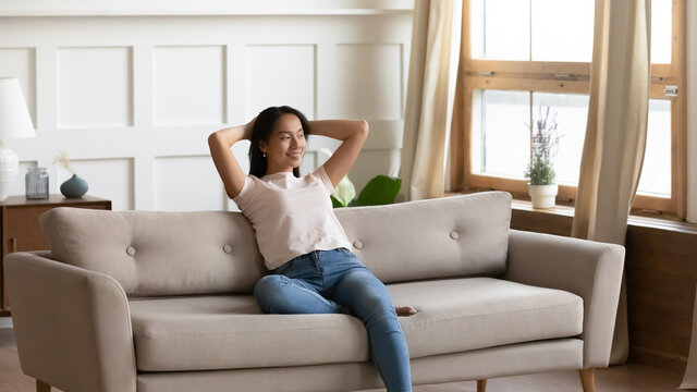 Satisfied dreamy young Asian woman relaxing on cozy couch in living room, smiling happy girl leaning back, stretching, planning future, dreaming, visualizing, enjoying lazy leisure time at home