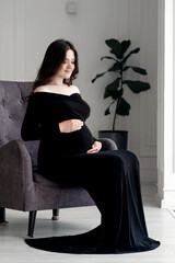 beautiful young pregnant woman in dark black dress sits on an armchair