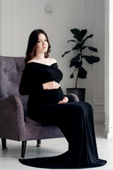 beautiful young pregnant woman in dark black dress sits on an armchair