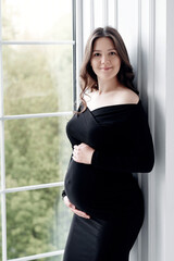 beautiful young pregnant woman in dark black dress stands near the window