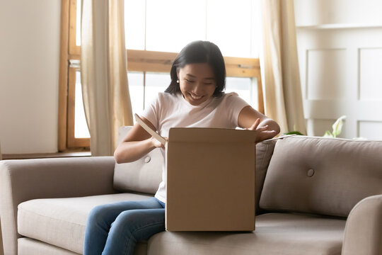 Smiling Young Asian Woman Opening Cardboard Box At Home, Sitting On Couch In Living Room, Satisfied Happy Girl Unpacking Parcel With Online Store Order Or Gift, Good Delivery Service Concept