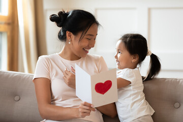 Smiling Asian young mother holding greeting card with red heart, sitting on couch at home, cute little girl daughter congratulating mum with mothers day or birthday, family celebration