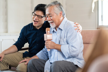Happy Asian family. Senior Asian father and Adult son singing karaoke on the sofa in the living...