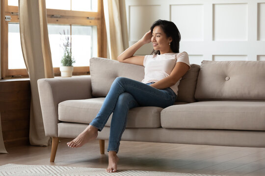 Smiling dreamy young Asian woman sitting on cozy couch in living room, looking to aside, thoughtful satisfied girl planning day, dreaming, visualizing future, enjoying leisure time at home