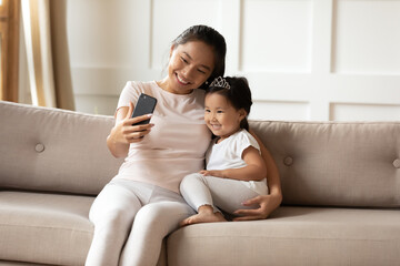 Smiling Asian mother and little daughter wearing princess diadem taking selfie together, happy young mum holding smartphone, hugging adorable toddler child girl, making video call, having fun