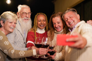 Happy senior people cheering with wine while taking a selfie with smartphone - Multiracial friends wearing warm christmas clothes