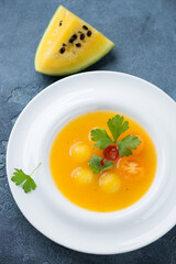 White plate with yellow watermelon and tomato gazpacho, elevated view on a blue stone background, vertical shot