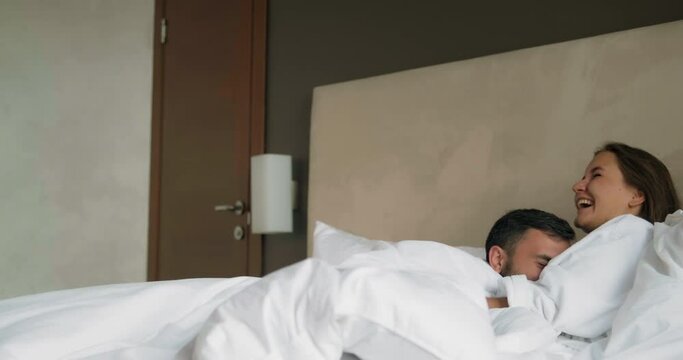 Joyful bearded man in bathrobe jumps to happy young woman and hugs lying on large bed in hotel room in morning at honey moon
