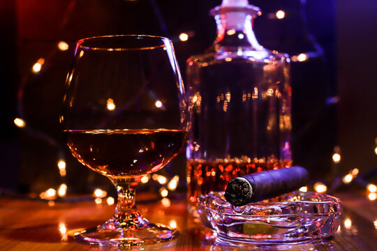 BOTTLE AND GLASS OF BORBON WITH CIGAR AND CHRISTMAS LIGHTS AND COLORED LIGHTS ON WOODEN TABLE AND DARK BACKGROUND