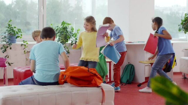 Medium shot of group of happy little schoolchildren running into classroom and sitting down to finish homework in their workbooks before lesson
