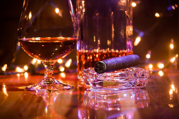 BOTTLE AND GLASS OF BORBON WITH CIGAR AND CHRISTMAS LIGHTS AND COLORED LIGHTS ON WOODEN TABLE AND DARK BACKGROUND