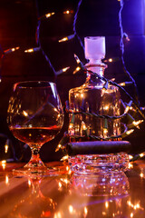 BOTTLE AND GLASS OF BORBON WITH CIGAR AND CHRISTMAS LIGHTS AND COLORED LIGHTS ON WOODEN TABLE AND DARK BACKGROUND